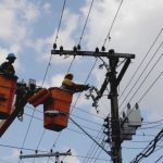 Técnicos em eletricidade trabalham em postes de energia, com céu azul e nuvens ao fundo.