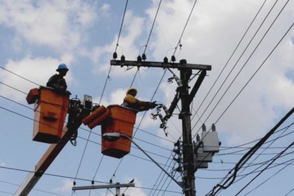Técnicos em eletricidade trabalham em postes de energia, com céu azul e nuvens ao fundo.