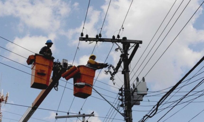 Técnicos em eletricidade trabalham em postes de energia, com céu azul e nuvens ao fundo.
