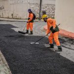 Trabalhadores asfaltando rua em Itapiranga, com uniforme laranja e ferramentas.