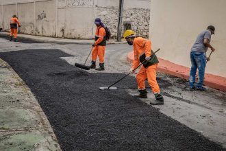 Trabalhadores asfaltando rua em Itapiranga, com uniforme laranja e ferramentas.