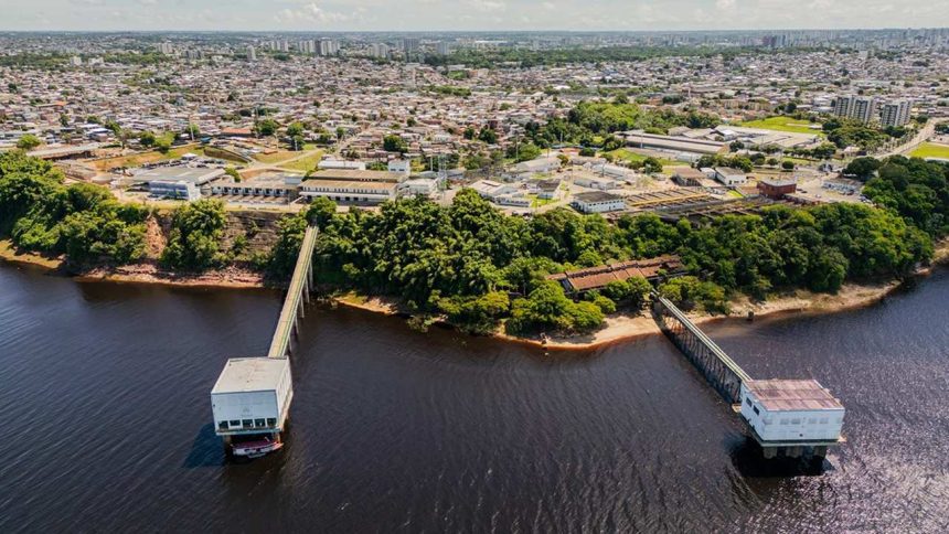 Vista aérea de estação de tratamento de água em Manaus, com cidade ao fundo e rio em primeiro plano.