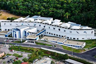 Vista aérea de um edifício branco moderno com detalhes em azul, cercado por vegetação exuberante. Se trata do prédio do Centro de Biotecnologia da Amazônia (CBA).