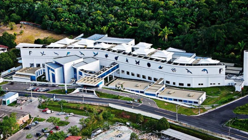 Vista aérea de um edifício branco moderno com detalhes em azul, cercado por vegetação exuberante. Se trata do prédio do Centro de Biotecnologia da Amazônia (CBA).