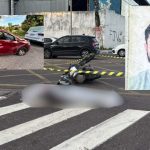 Acidente de trânsito com carro vermelho e moto, com corpo no chão e foto de homem.