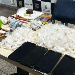 Apreensão de drogas e objetos em uma mesa, incluindo celulares, dinheiro e embalagens de entorpecentes.