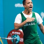 Atleta brasileiro comemorando vitória no levantamento de peso, vestindo uniforme do Brasil.