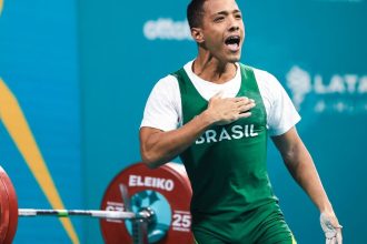 Atleta brasileiro comemorando vitória no levantamento de peso, vestindo uniforme do Brasil.