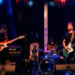 Banda de rock tocando no palco, guitarrista, baixista e baterista em ação.