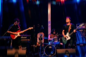 Banda de rock tocando no palco, guitarrista, baixista e baterista em ação.