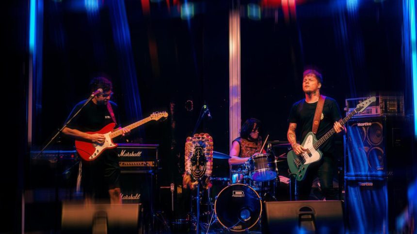 Banda de rock tocando no palco, guitarrista, baixista e baterista em ação.