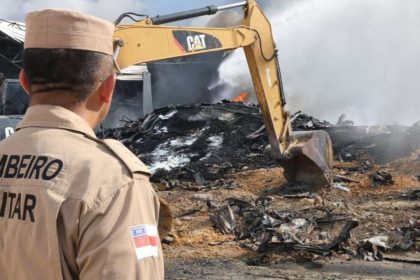 Bombeiro observa destroços de incêndio com máquina, fumaça e chamas visíveis.