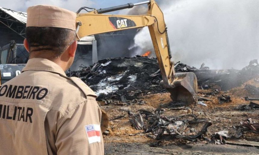 Bombeiro observa destroços de incêndio com máquina, fumaça e chamas visíveis.