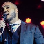 Cantor Belo cantando em um palco, segurando um microfone. Ele usa um terno e gravata.