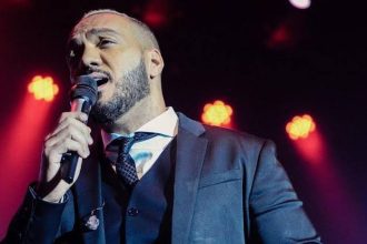 Cantor Belo cantando em um palco, segurando um microfone. Ele usa um terno e gravata.