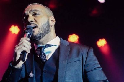 Cantor Belo cantando em um palco, segurando um microfone. Ele usa um terno e gravata.