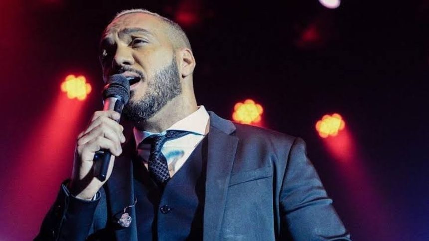 Cantor Belo cantando em um palco, segurando um microfone. Ele usa um terno e gravata.