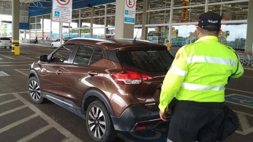 Carro marrom em vaga especial levando multa para deficientes com guarda de trânsito ao lado.