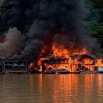 Casas flutuantes em chamas em rio, com fumaça escura e vegetação ao fundo.