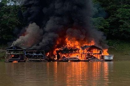Casas flutuantes em chamas em rio, com fumaça escura e vegetação ao fundo.