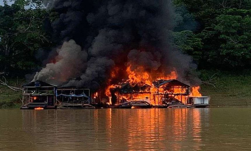 Casas flutuantes em chamas em rio, com fumaça escura e vegetação ao fundo.