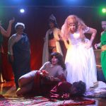 Cena da peça 'Medéia' em versão cômica e ousada, com elenco em figurinos teatrais e iluminação dramática.