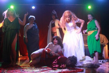 Cena da peça 'Medéia' em versão cômica e ousada, com elenco em figurinos teatrais e iluminação dramática.