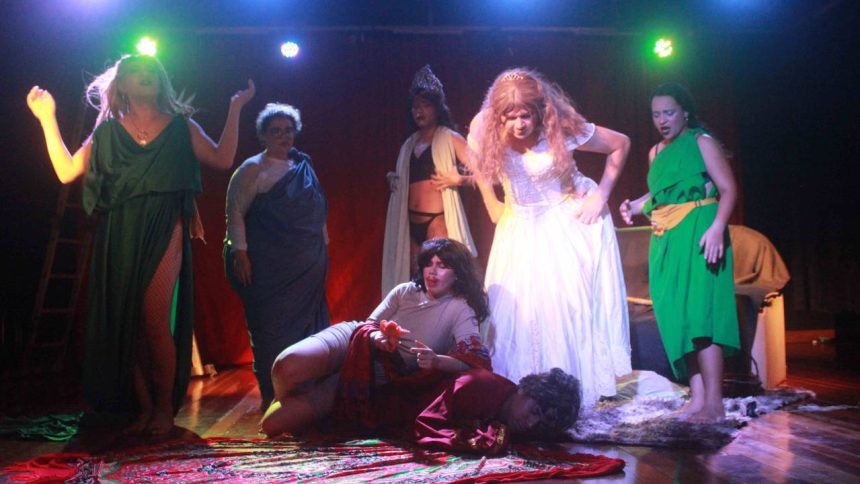 Cena da peça 'Medéia' em versão cômica e ousada, com elenco em figurinos teatrais e iluminação dramática.