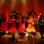 Cena de dança vibrante no palco do Teatro Amazonas, com figurinos coloridos e luzes dramáticas.