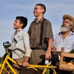 Cena de filme com três homens sorrindo ao lado de uma bicicleta amarela