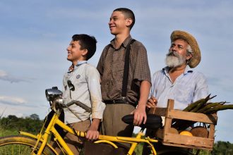 Cena de filme com três homens sorrindo ao lado de uma bicicleta amarela