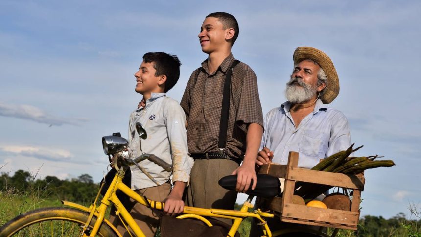 Cena de filme com três homens sorrindo ao lado de uma bicicleta amarela