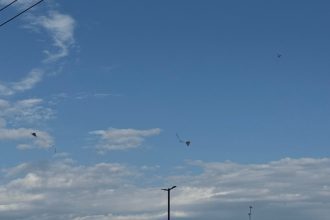Céu azul de calor com nuvens e pipas voando, criando um cenário de dia ensolarado.