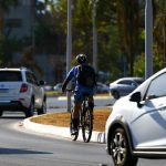 Ciclista pedalando em via urbana, com carros e árvores ao fundo, em dia ensolarado.