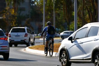 Ciclista pedalando em via urbana, com carros e árvores ao fundo, em dia ensolarado.