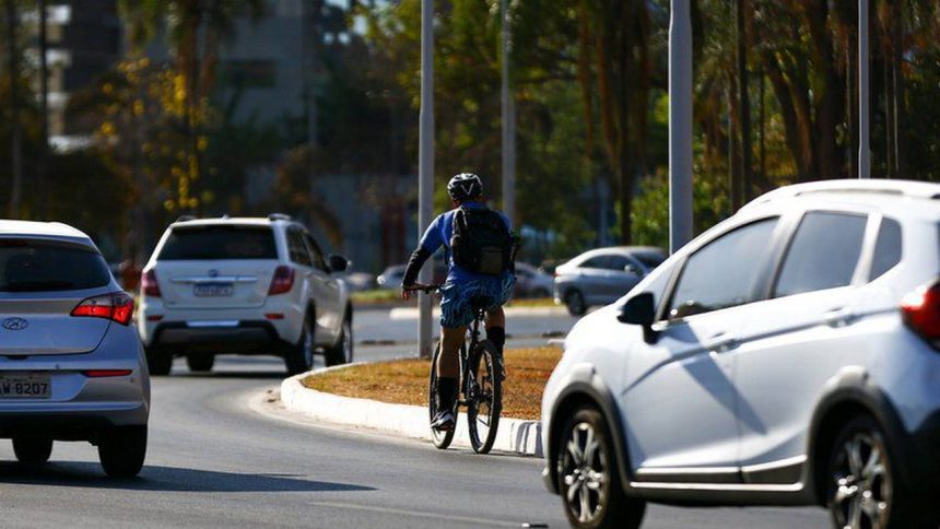 Ciclista pedalando em via urbana, com carros e árvores ao fundo, em dia ensolarado.