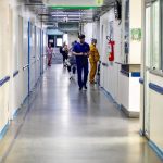 Corredor de hospital vazio, com funcionários caminhando e luz natural.