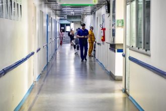 Corredor de hospital vazio, com funcionários caminhando e luz natural.