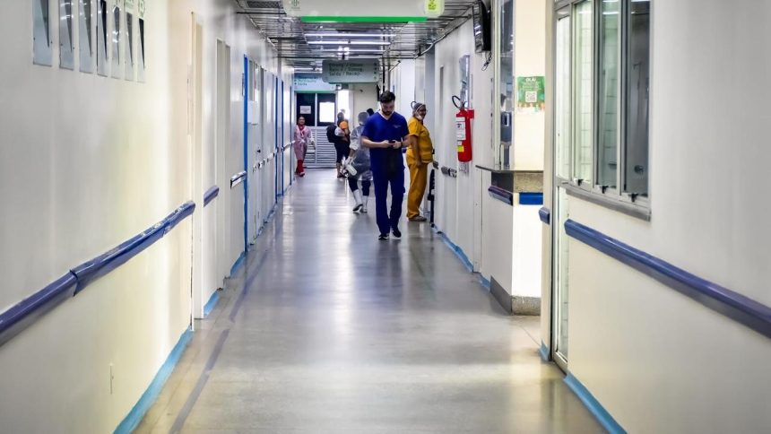 Corredor de hospital vazio, com funcionários caminhando e luz natural.