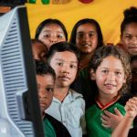 Escolas conectadas: Crianças sorrindo em volta de um monitor de computador, indicando um ambiente educacional.