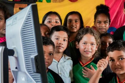 Escolas conectadas: Crianças sorrindo em volta de um monitor de computador, indicando um ambiente educacional.