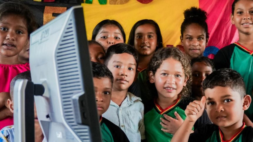 Escolas conectadas: Crianças sorrindo em volta de um monitor de computador, indicando um ambiente educacional.