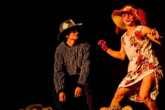 Dois atores em figurino de peça teatral, um com chapéu e o outro com vestido floral, em palco escuro.