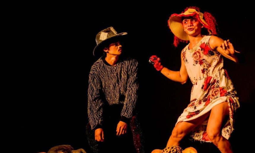Dois atores em figurino de peça teatral, um com chapéu e o outro com vestido floral, em palco escuro.