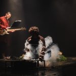 Dois atores em um palco escuro com fumaça e instrumentos musicais.