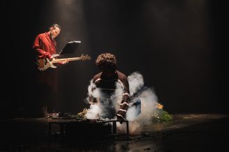 Dois atores em um palco escuro com fumaça e instrumentos musicais.