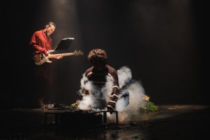 Dois atores em um palco escuro com fumaça e instrumentos musicais.