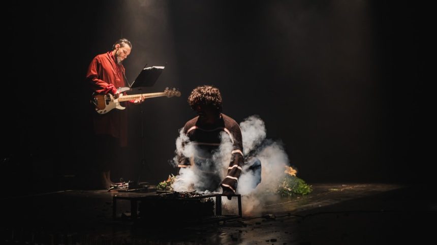 Dois atores em um palco escuro com fumaça e instrumentos musicais.
