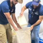 Dois homens em Parintis transferindo um bloco de produto embalado para um saco grande dentro de uma sala.