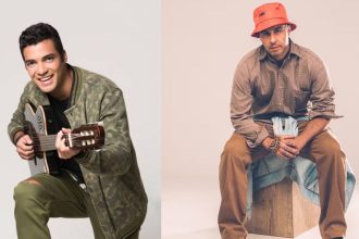 Dois homens, um tocando violão e outro sentado em um banco, com boné.